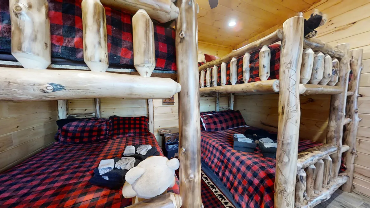 A cozy bunk room features two Queen-over-Queen bunk beds with plaid bedding, wooden log accents, and a stuffed teddy bear. The room feels playful and inviting, ideal for families or groups sharing space and creating memories.