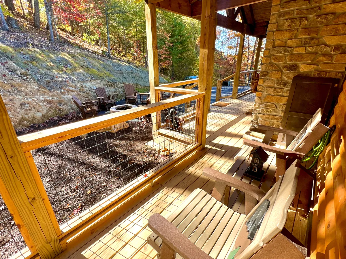 A spacious wooden deck bordered by a rustic stone wall, complete with inviting outdoor seating and a modern fire pit. The deck overlooks serene, tree-filled surroundings, capturing the tranquil essence of mountain living. The cozy atmosphere is enhanced by the natural beauty, perfect for gatherings or quiet moments.