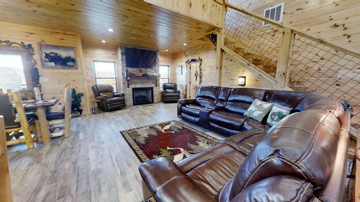 A spacious open-concept living area featuring leather couches arranged around a cozy fireplace. The dining area is visible in the background, and large windows frame scenic mountain views, allowing natural light to pour in. The room exudes warmth and is ideal for gatherings, relaxing, or sharing a meal.