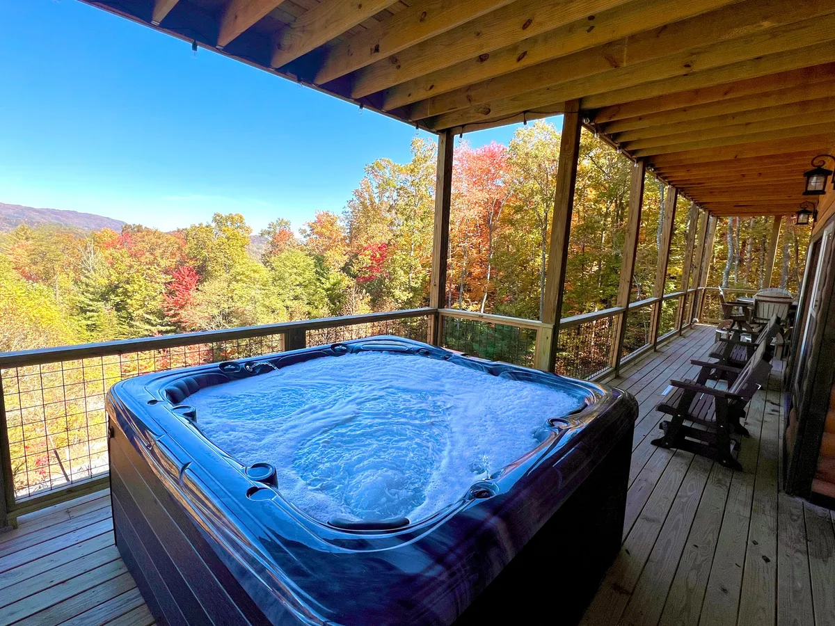 A bubbling hot tub sits on a wooden deck, overlooking a stunning view of mountains covered in fall foliage. The setting is serene and inviting, with space for several people to unwind and soak in the natural beauty that surrounds the lodge.