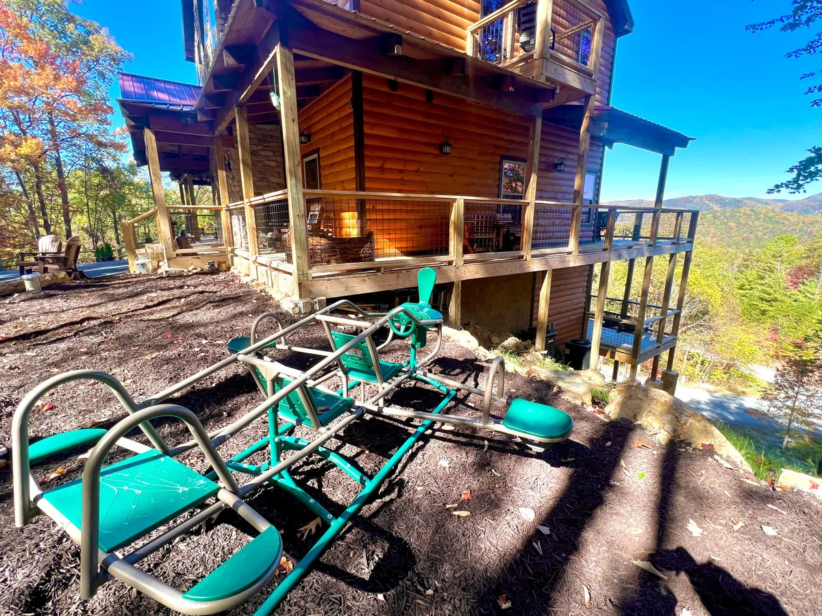 A wooden deck features a well-equipped outdoor play area with playground equipment for children, overlooking scenic mountain views. The space combines areas for both relaxation and play, set against the majestic landscape.