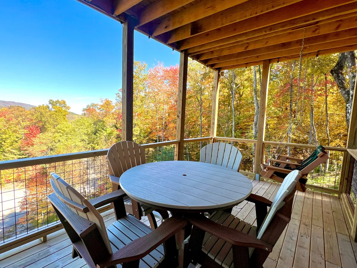 A wooden outdoor table surrounded by classic Adirondack chairs is set on a deck, framed by the vibrant colors of autumn foliage. The scene is tranquil and welcoming, ideal for outdoor dining, relaxing with a book, or socializing with family and friends while immersed in the beauty of the Smoky Mountains.