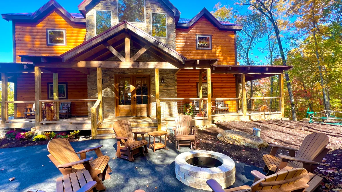 The image showcases the exterior of a large, inviting wooden cabin surrounded by lush trees. A glowing fire pit, encircled by Adirondack chairs, sits in the foreground, beckoning guests to gather and unwind. The porch provides sunset views, hinting at tranquil evenings spent outdoors, while the natural setting suggests peaceful nature walks and stargazing under Tennessee skies.