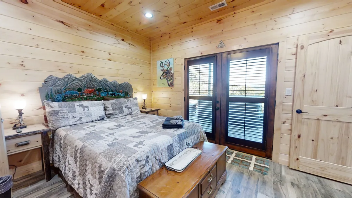 A bedroom with a decorative headboard and soft bedding opens directly onto a private deck through double doors. A nightstand with a lamp and a cozy rug complete the inviting look, with natural light pouring in and a hint of the outdoors visible beyond the doors.