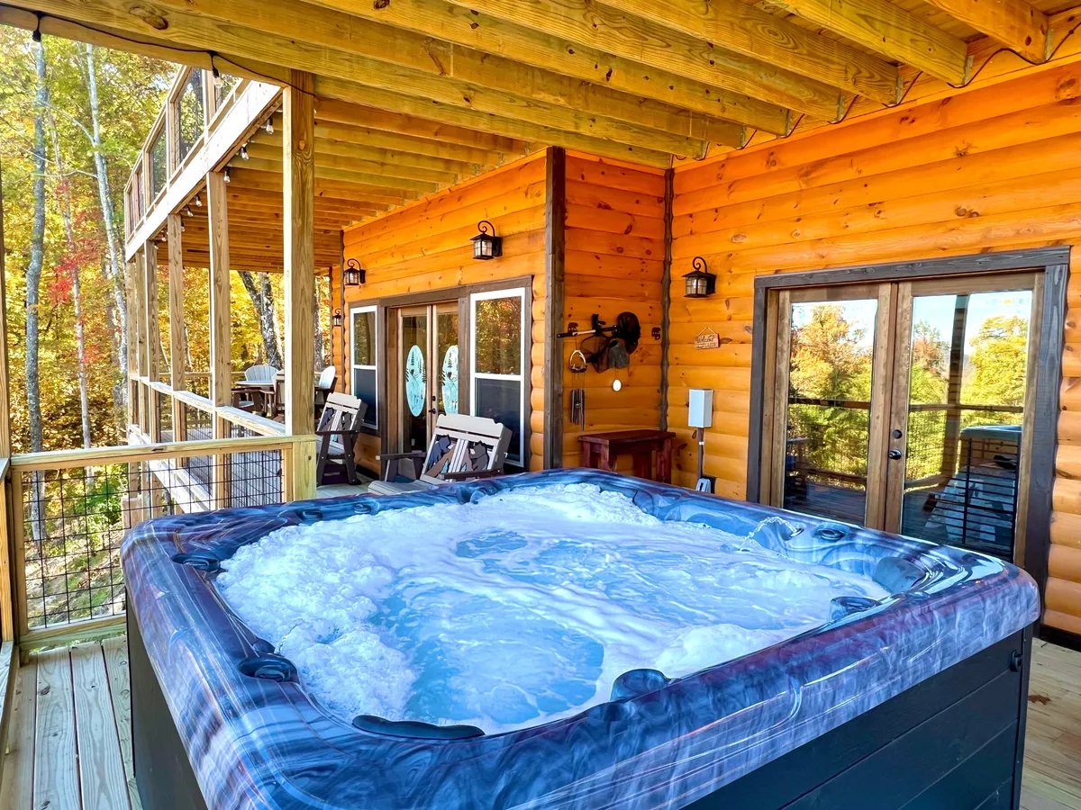 A bubbling hot tub sits on a wooden deck, backed by the log cabin structure and surrounded by lush trees. The tranquil setting captures a sense of comfort and relaxation, inviting guests to enjoy mountain air from the privacy of the tub.