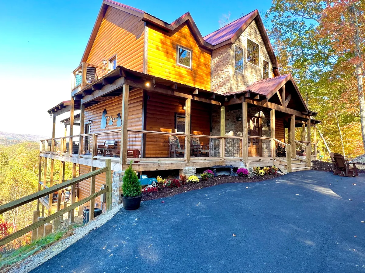 A spacious wooden deck adorned with colorful flowers overlooks a stunning mountain view. The stone facade of the lodge and abundant greenery evoke a welcoming atmosphere, perfect for gatherings, sunrise coffees, or nature appreciation in spring and summer.