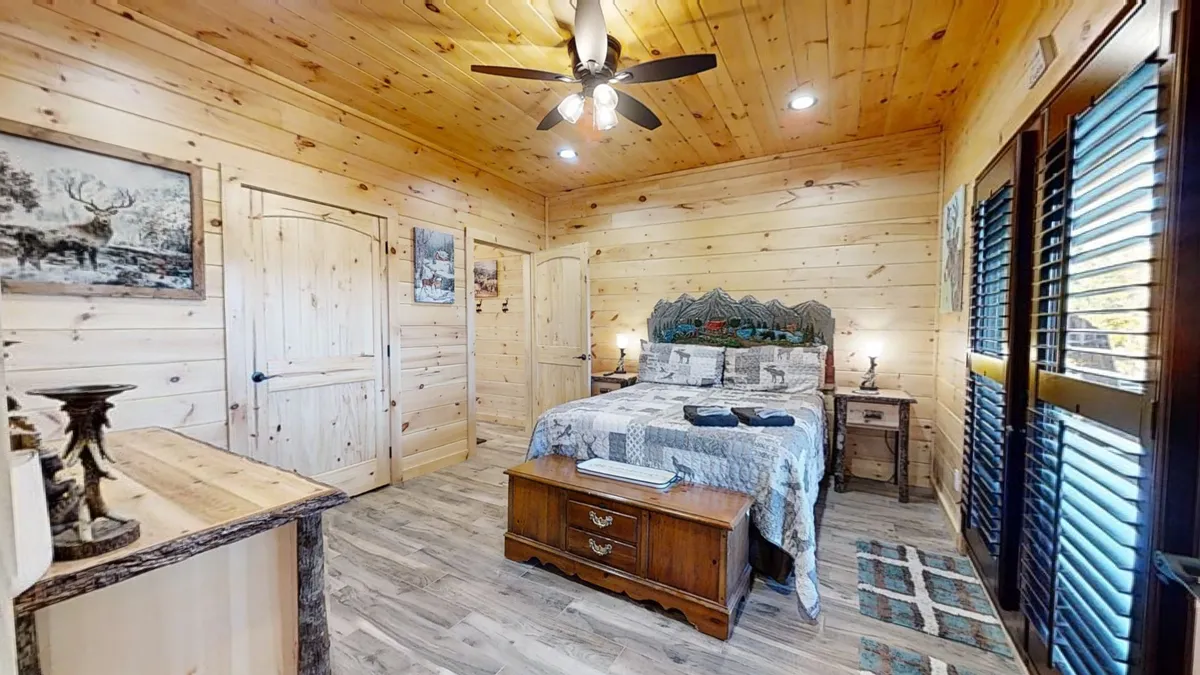 A warmly lit bedroom showcases a wooden bed frame adorned with plaid bedding, a wooden nightstand, and rustic artwork on the walls. Natural light streams into the room, highlighting the inviting and tranquil ambiance, perfect for a restful night's sleep.